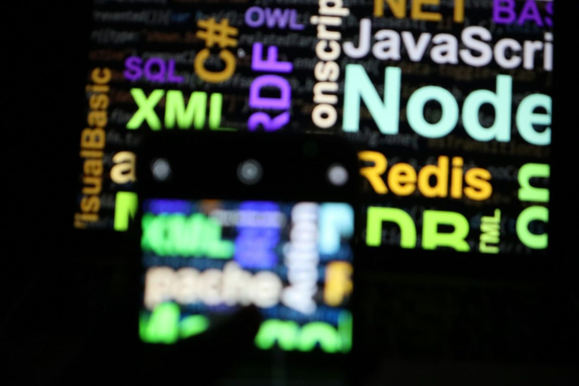 Photographie en faible luminosité d'un écran d'ordinateur flou affichant des noms de technologies de programmation comme "JavaScript" et "Node.js", avec un smartphone au premier plan affichant des termes similaires, illustrant l'importance des outils numériques modernes et des outils IA essentiels pour les professionnels et particuliers.