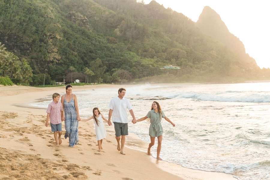 Une famille de quatre personnes, parents et deux enfants, marche main dans la main sur une plage de sable au coucher du soleil, avec des montagnes verdoyantes en arrière-plan, lors d'un voyage en famille.